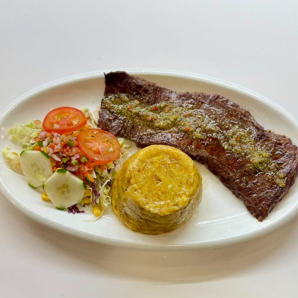 Grilled steak topped with green herb sauce, served with a fresh salad and golden fried plantain on a white plate.