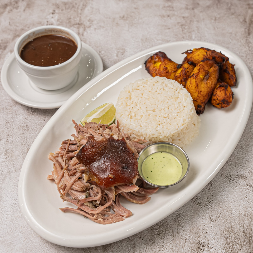 Plate of shredded roasted pork, white rice, fried plantains, lime, dipping sauce, and a cup of dark soup.