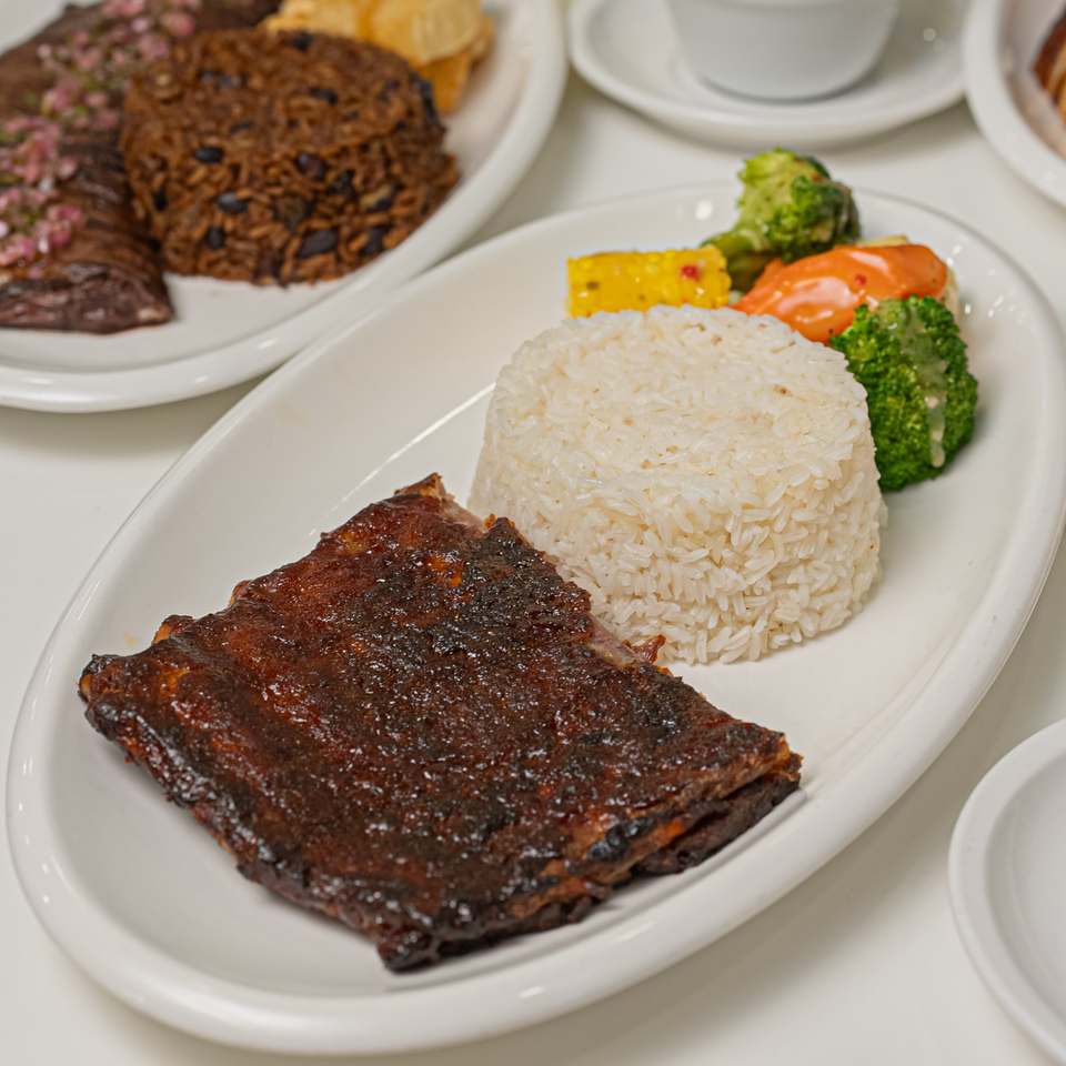 Glazed barbecue ribs served with a mound of white rice, broccoli, carrots, and corn on a white platter.