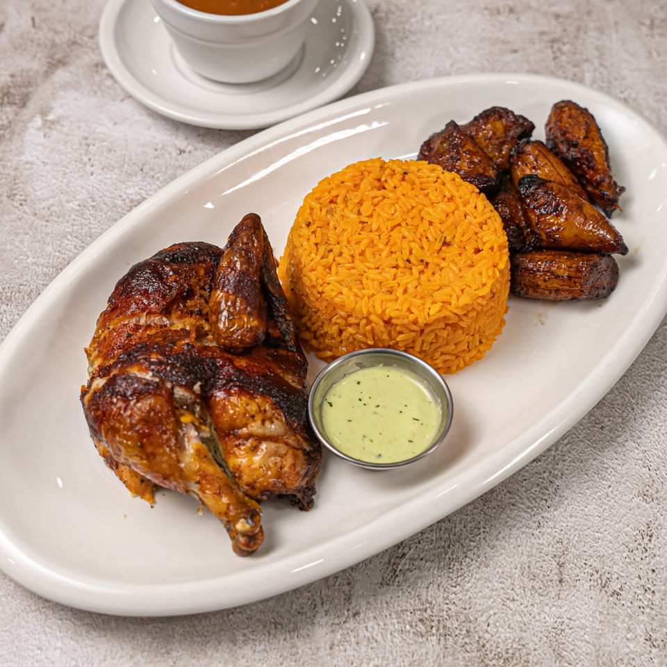 Grilled chicken with orange rice, roasted plantains, and creamy green dipping sauce on a white platter.