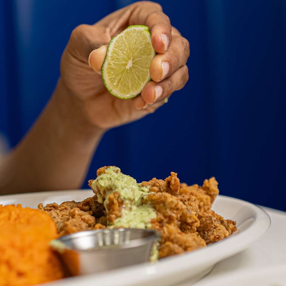 Hand holds a lime wedge over crispy fried food topped with green sauce, served with a dipping cup and orange side.